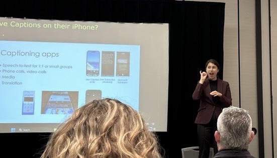A presenter addressing an audience beside a slide about captioning apps